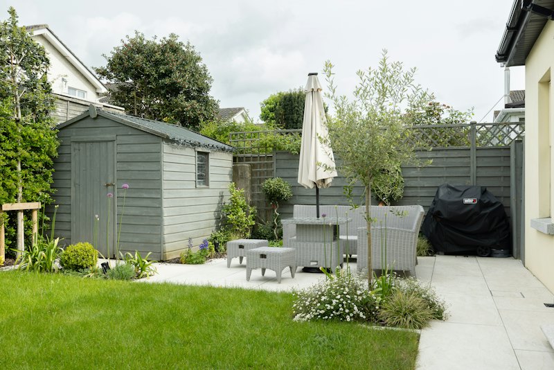 A back garden with a pale stone patio, lawn, shed and outdoor seating